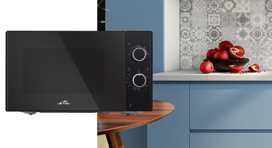 Czarna kuchenka mikrofalowa z dwoma pokrętłami i logo \'eta\' na drzwiczkach. W tle widoczna jest kuchnia z niebieskimi szafkami i misą z granatami na blacie.