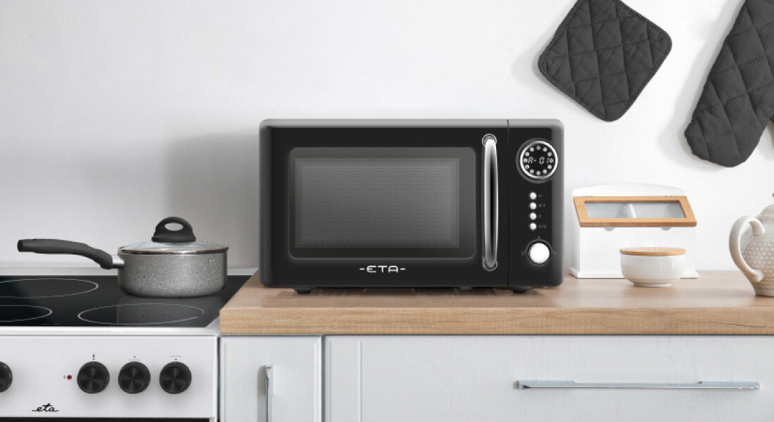 Kuchenka mikrofalowa ETA Storio ETA321090020 Zdjęcie przedstawia czarną kuchenkę mikrofalową ETA ustawioną na drewnianym blacie w jasnej kuchni, obok płyty grzewczej i garnka. Minimalistyczna aranżacja podkreśla elegancki design urządzenia
