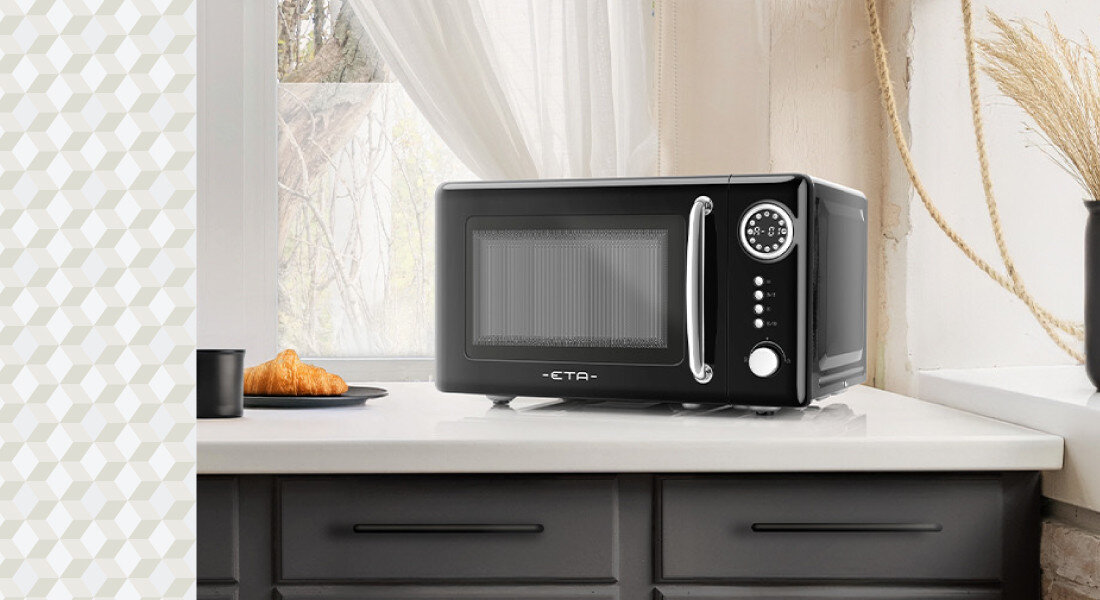 Kuchenka mikrofalowa ETA Storio ETA321090020 Zdjęcie ukazuje kuchenkę mikrofalową ETA ustawioną na jasnym blacie przy oknie, obok talerza z rogalikiem. Naturalne światło podkreśla błyszczący front i kompaktową formę urządzenia mikrofalówka retro czarna obudowa styl kuchnia design funkcjonalność