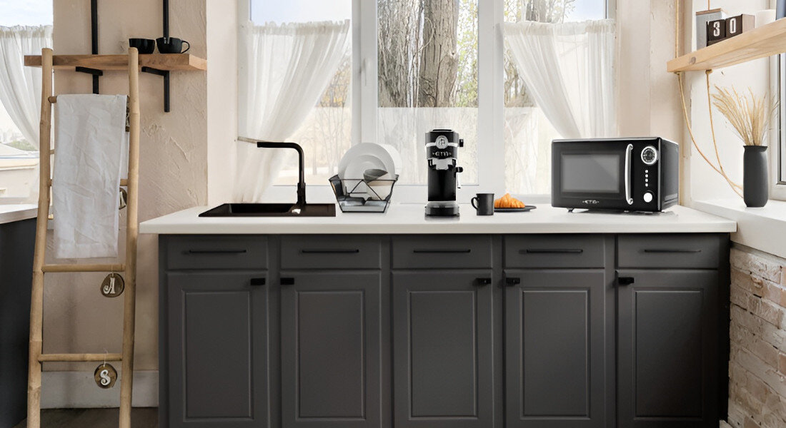 Kuchenka mikrofalowa ETA Storio ETA321090020 Zdjęcie ukazuje kuchenkę mikrofalową ETA w szerokiej aranżacji kuchennej, ustawioną obok ekspresu do kawy i zlewu. Kompozycja podkreśla dopasowanie urządzenia do wnętrza pojemność 20l talerz obrotowy podświetlenie wnętrza kompaktowe wymiary wygoda