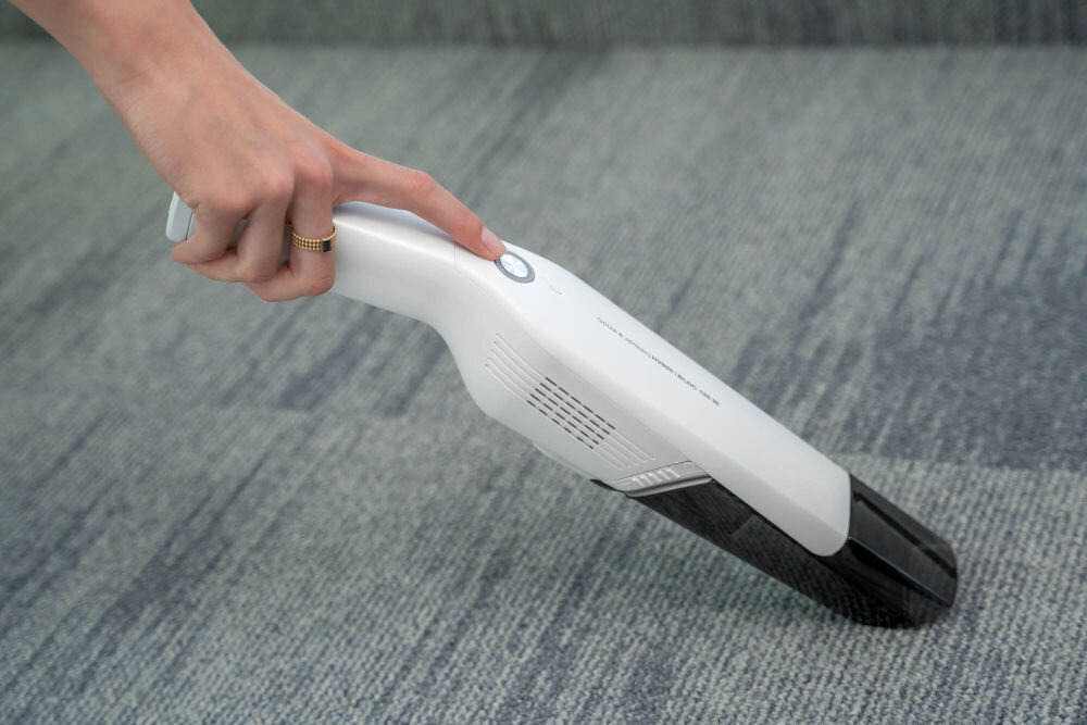 Odkurzacz ręczny GÖTZE & JENSEN HV800 Dłoń z pierścionkiem naciska przycisk zasilania na górze białego, ręcznego odkurzacza Gotze & Jensen HV800, trzymając go nad szarym, wykładzinowym podłożem