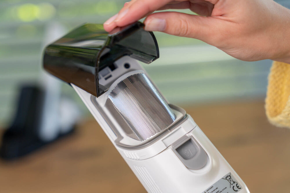 Odkurzacz ręczny GÖTZE & JENSEN HV800 Zbiornik na kurz ręcznego odkurzacza Gotze & Jensen HV800 jest otwarty, ukazując metalowy filtr siatkowy wewnątrz. Ręka unosi czarną pokrywę