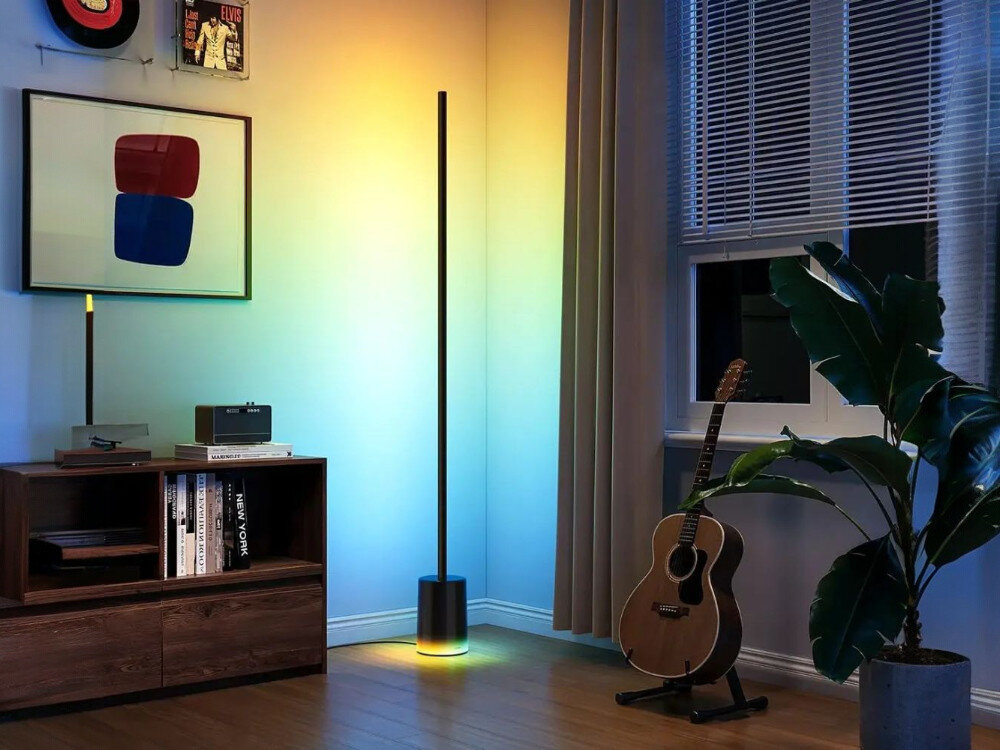 Lampa podłogowa GOVEE H607C, design, słup, podstawa, aranżacja, Smukła czarna lampa podłogowa Govee w rogu salonu emituje tęczowe gradienty RGB (żółty, pomarańczowy, niebieski, zielony), obok gitary, książek, rośliny i głośnika, tworząc dynamiczną atmosferę wieczorem z zasłonami i plakatami na ścianach.