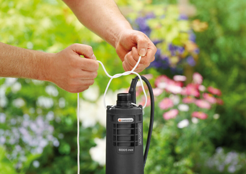 Osoba trzyma czarną pompę do wody z widocznym logo \'GARDENA\' i napisem \'6000/5 inox\'. W tle znajduje się rozmyty ogród z kwiatami.