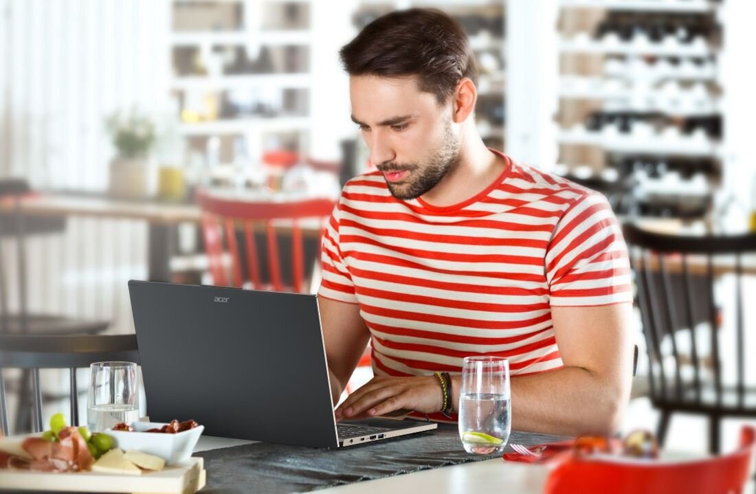 Osoba siedzi przy stole, używając laptopa z widocznym logo Acer. Na stole znajdują się szklanki z wodą i talerze z jedzeniem.