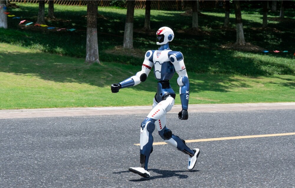 Robot humanoidalny idzie szerokim krokiem po czarnej, asfaltowej drodze, w tle widać rzędy drzew i trawnik