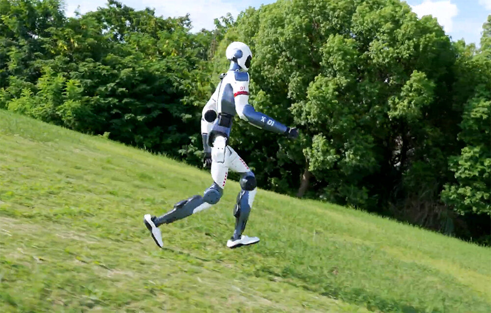 Robot humanoidalny w trakcie biegu po zielonym, trawiastym zboczu, w tle widać gęstą ścianę drzew.