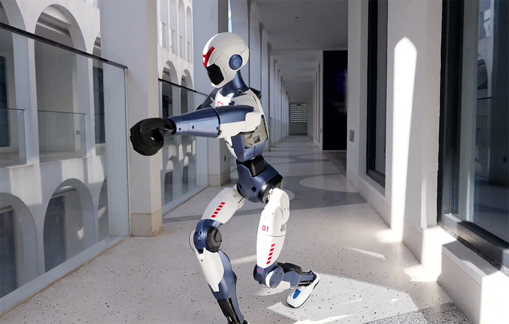 Robot humanoidalny uchwycony w półprzysiadzie wewnątrz długiego, jasnego korytarza z widocznymi łukami.