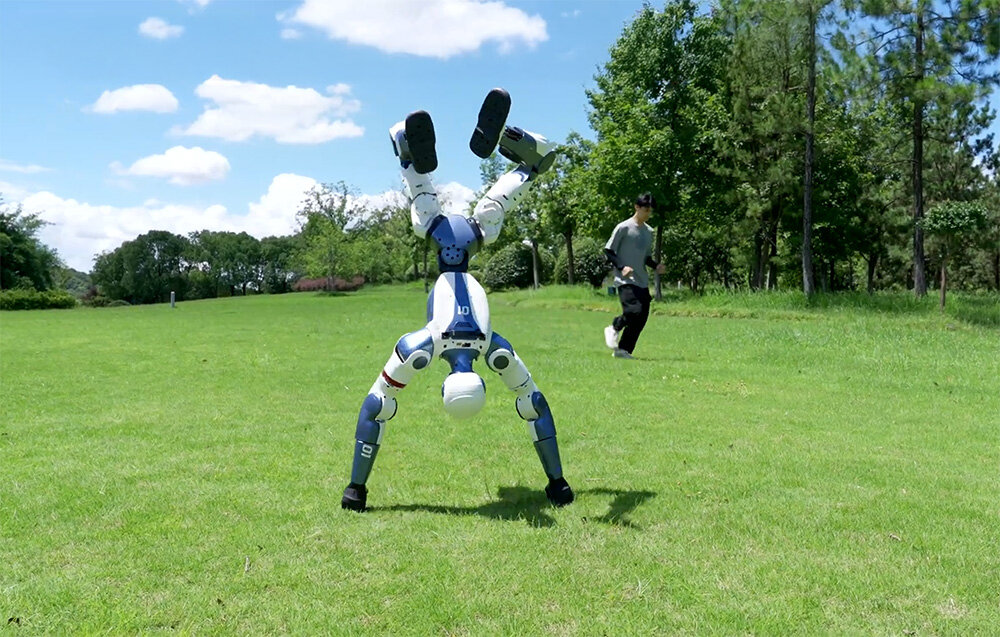 Robot humanoidalny stoi na rękach na zielonej trawie, w tle po prawej stronie biegnie mężczyzna