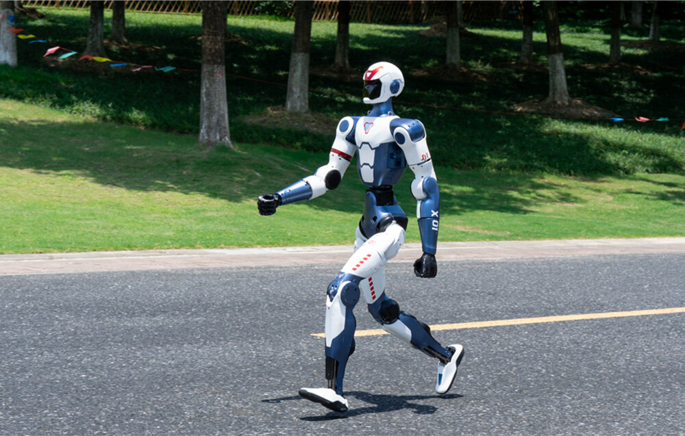 Robot humanoidalny idzie szerokim krokiem po czarnej, asfaltowej drodze, w tle widać drzewa.