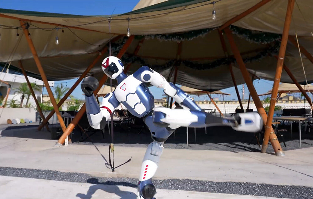 Robot humanoidalny stoi na jednej nodze, wykonując wysokie kopnięcie przed dużym namiotem.