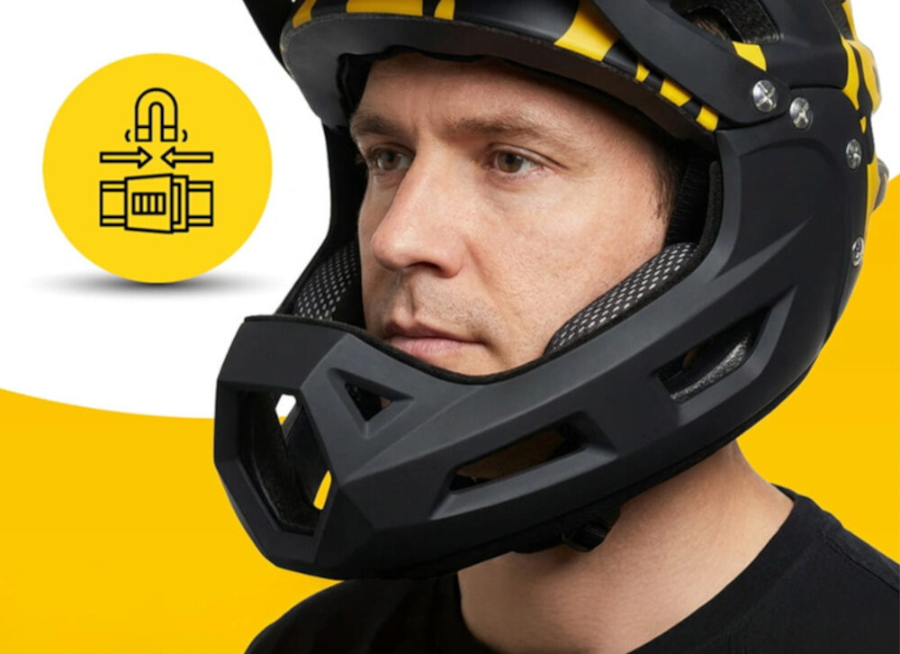 Osoba nosi czarno-żółty kask typu full face z odkręcaną szczęką. Obok znajduje się żółte koło z czarnym symbolem przedstawiającym mechanizm odkręcania.