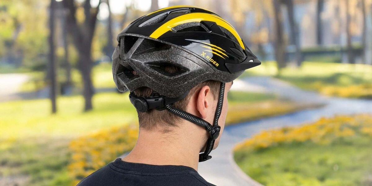 Osoba nosi czarno-żółty kask rowerowy z napisem \'XRIDER\' na tle parku. Kask ma aerodynamiczny kształt i regulowany pasek pod brodą.