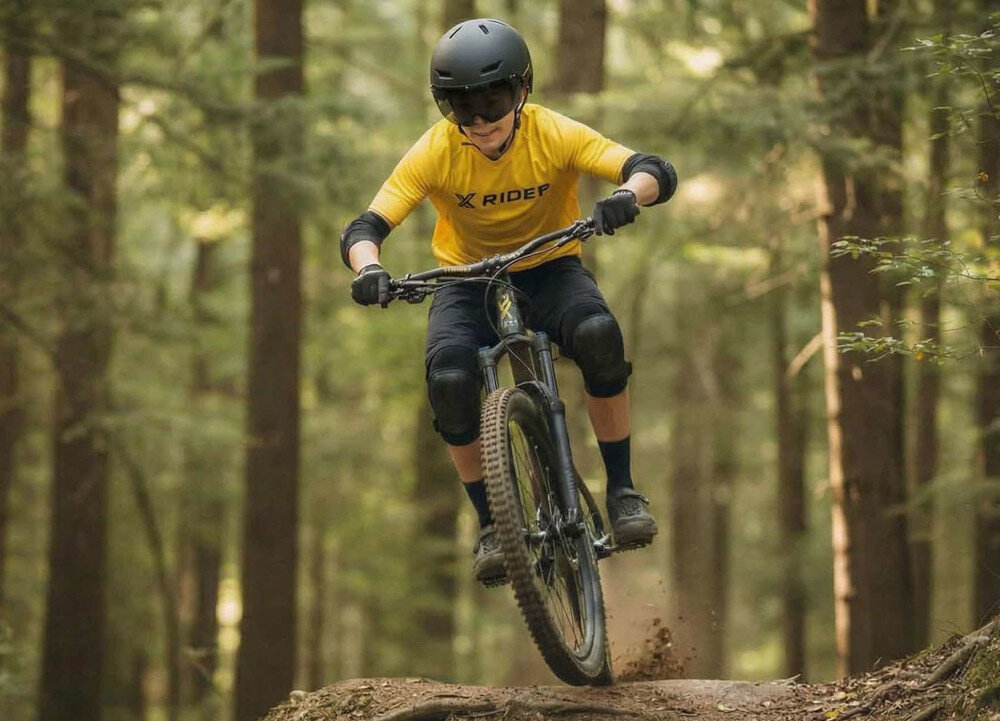 Osoba na rowerze górskim w lesie, ubrana w żółtą koszulkę z napisem \'X RIDER\' oraz czarny kask. W tle widoczne są drzewa i leśna ścieżka.