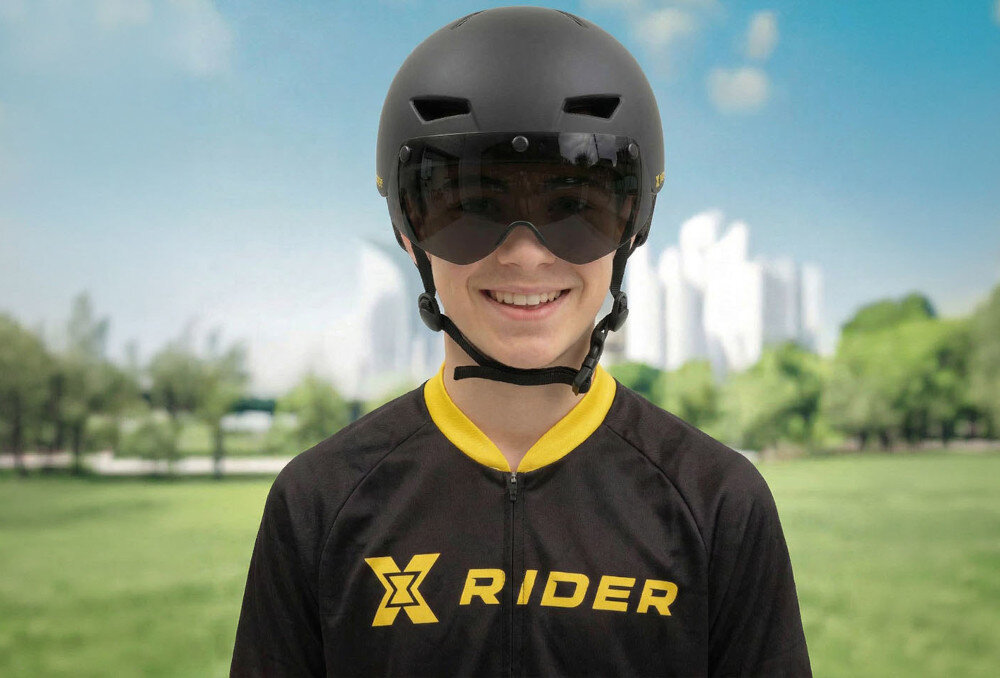 Osoba nosi czarny kask z przyciemnianą osłoną, ubrana w czarną koszulkę z żółtym logo \'XRIDER\'. W tle widać rozmyty krajobraz z drzewami i budynkami.