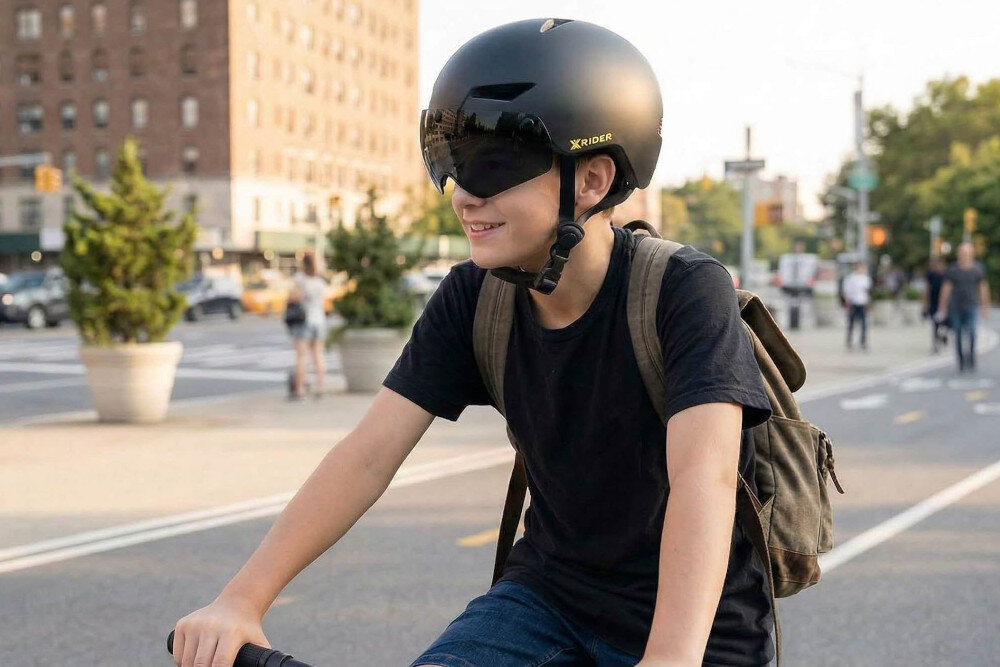 Osoba na rowerze nosi czarny kask z napisem \'XRIDER\'. W tle widać miejską ulicę z budynkami i roślinnością.