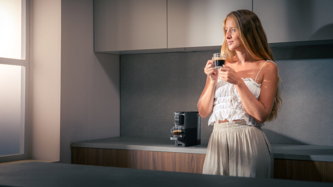 Kobieta stoi w kuchni, trzymając filiżankę kawy. Na blacie znajduje się ekspres do kawy.
