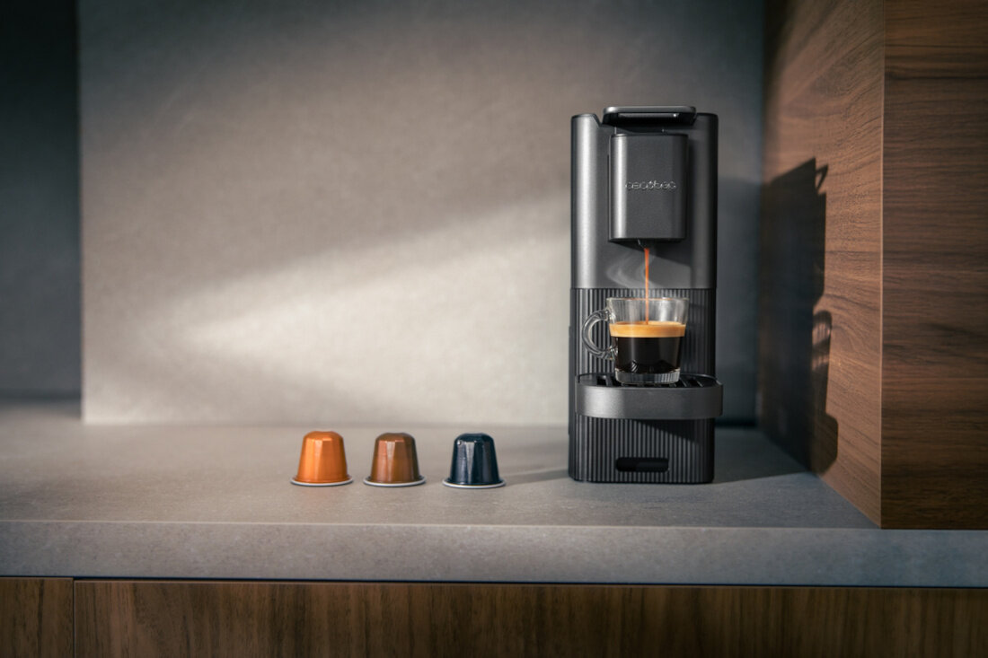 Na blacie kuchennym stoi czarny ekspres do kawy z filiżanką, z której leje się kawa. Obok ekspresu znajdują się trzy kolorowe kapsułki do kawy.