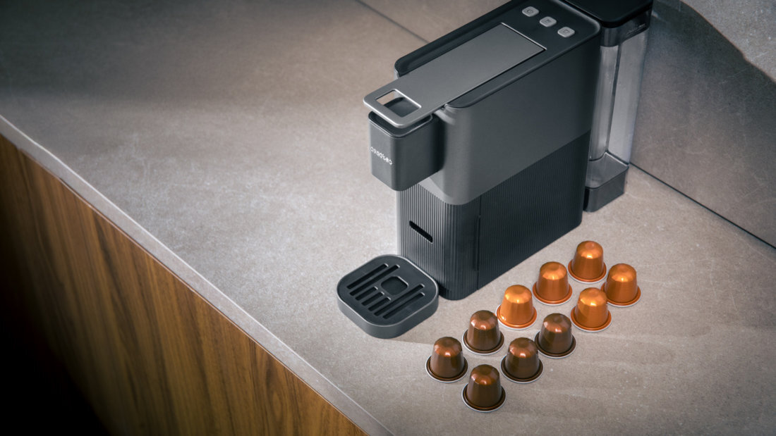 Na blacie kuchennym stoi czarny ekspres do kawy z otwartą tacką ociekową. Obok ekspresu leży dziewięć kapsułek do kawy w kolorze brązowym.