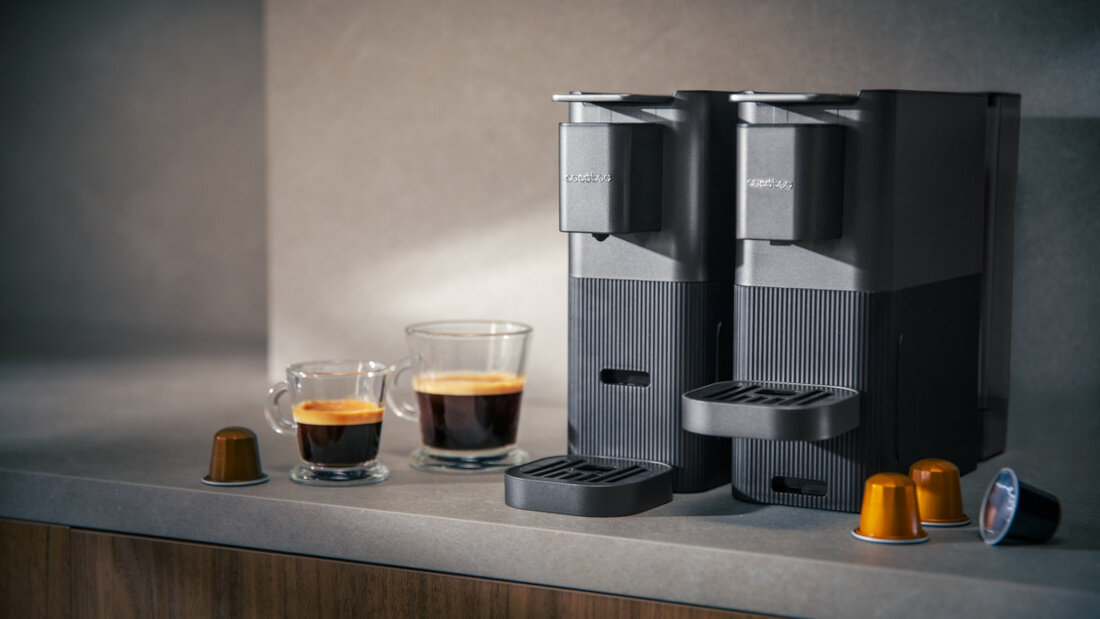 Na blacie kuchennym stoją dwa czarne ekspresy do kawy z logo \'cecotec\', obok nich znajdują się dwie filiżanki z kawą oraz kilka kapsułek do kawy.