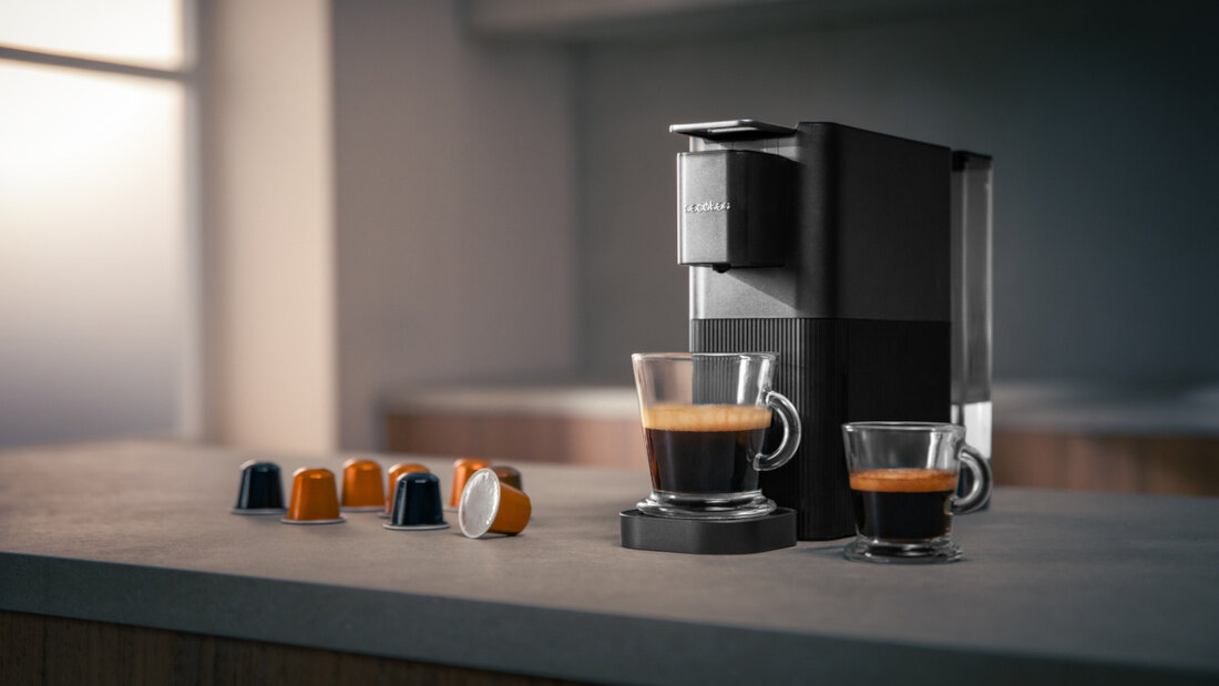 Na blacie kuchennym stoi czarny ekspres do kawy z logo \'CECOTEC\', obok którego znajdują się filiżanki z kawą i kilka kapsułek w różnych kolorach.