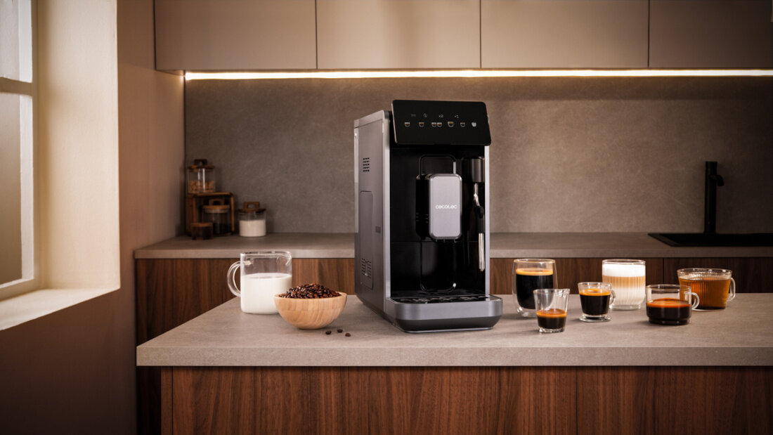 Na blacie kuchennym stoi ekspres do kawy z widocznym logo \'CECOTEC\'. Obok ekspresu znajdują się filiżanki z różnymi rodzajami kawy oraz dzbanek z mlekiem i pojemnik z ziarnami kawy.