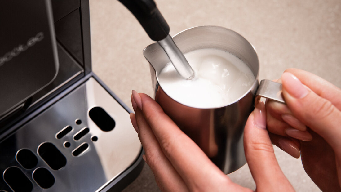 Osoba trzyma metalowy dzbanek z mlekiem, pod którym znajduje się dysza parowa ekspresu do kawy. Na ekspresie widoczne jest logo \'cecotec\'.