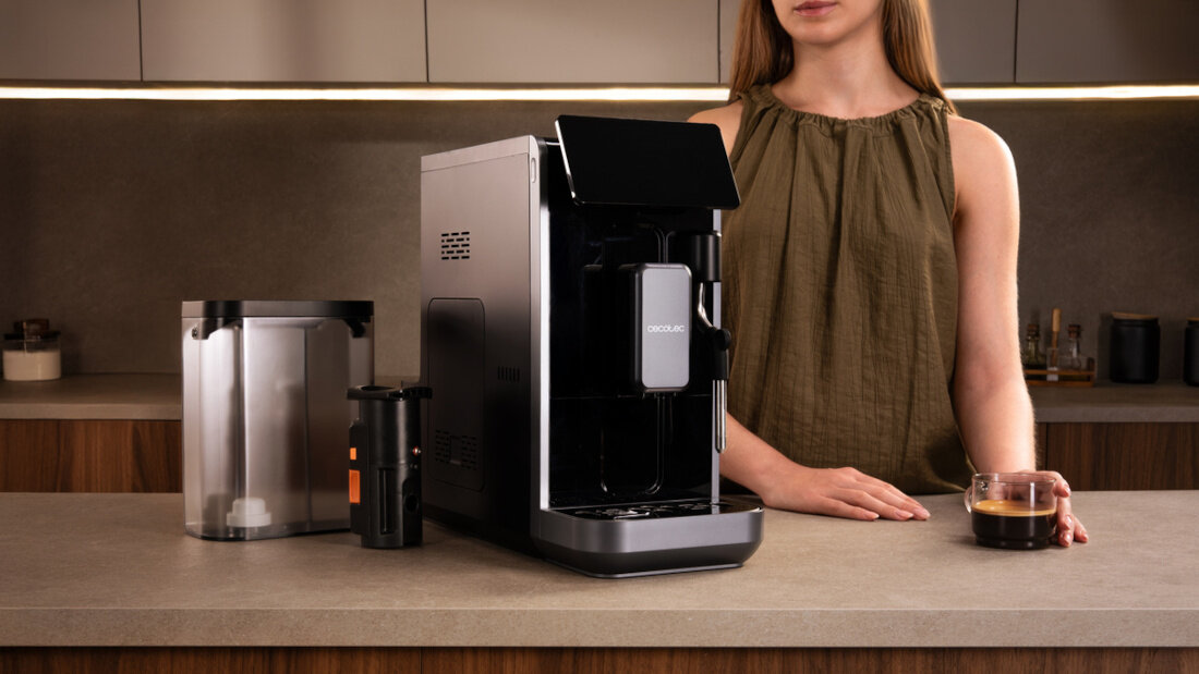 Na blacie kuchennym stoi ekspres do kawy z widocznym logo \'CECOTEC\', obok znajduje się pojemnik na wodę i filiżanka z kawą. W tle widać osobę w zielonej bluzce.