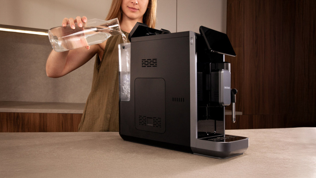 Osoba nalewa wodę z karafki do zbiornika ekspresu do kawy. Ekspres jest czarny z widocznym napisem \'CECOTEC\'.