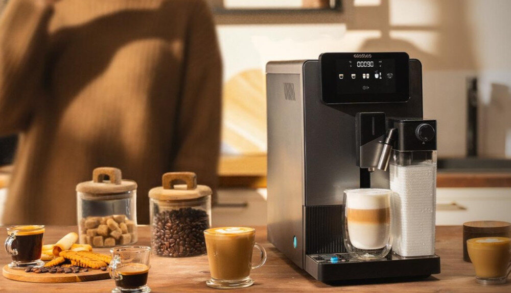 Na blacie kuchennym stoi ekspres do kawy z logo \'cecotec\', obok którego znajdują się filiżanki z kawą i słoiki z cukrem oraz ziarnami kawy. W tle widoczna jest osoba w swetrze.