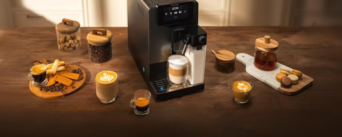 Na drewnianym stole znajduje się ekspres do kawy z wyświetlaczem, obok którego stoją filiżanki z kawą i słoiki z różnymi produktami spożywczymi. Ekspres przygotowuje kawę z mleczną pianką.