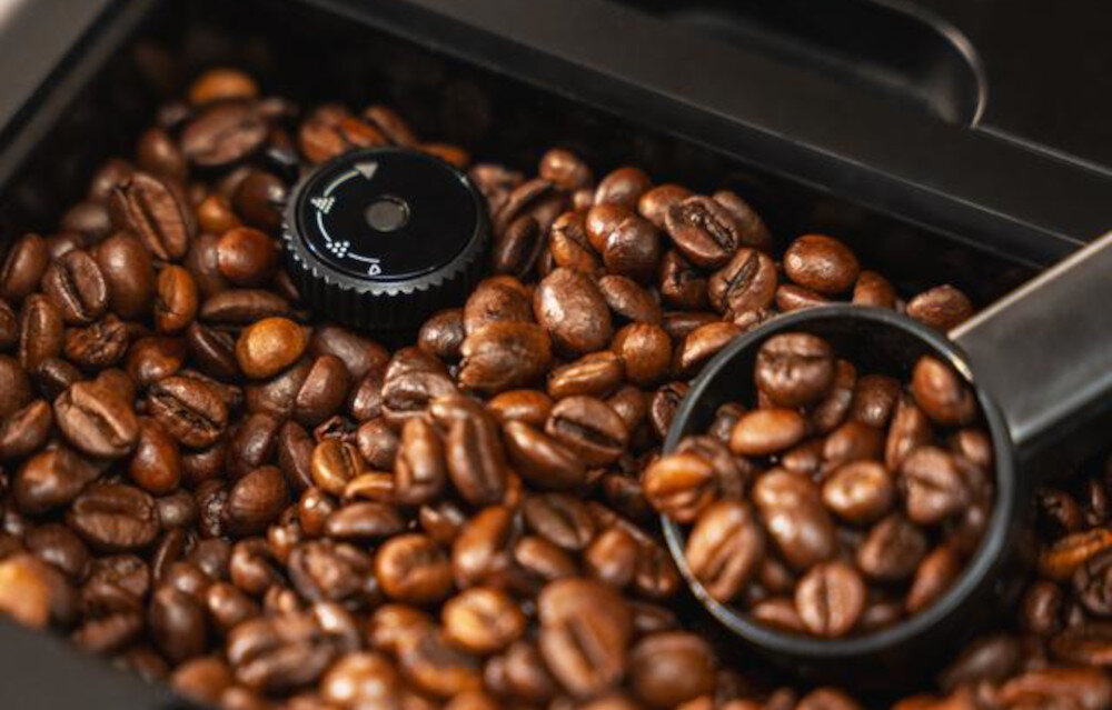 Zbliżenie na ziarna kawy w pojemniku z czarnym pokrętłem i miarką. Pokrętło ma oznaczenia w kształcie strzałek i symbolu filiżanki.