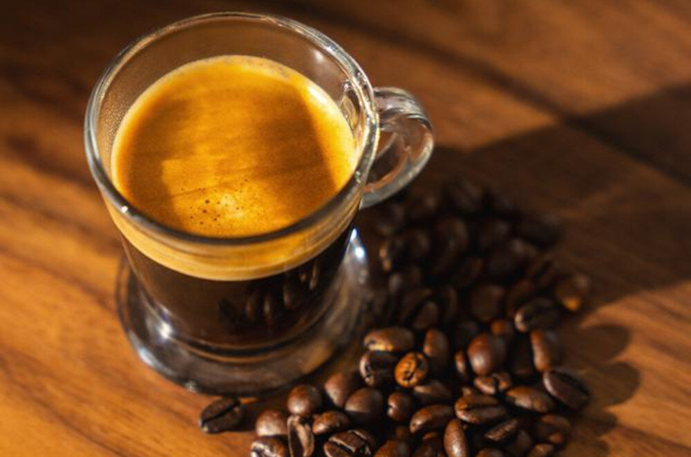 Szklanka espresso z kremową pianką stoi na drewnianym blacie, otoczona ziarnami kawy.