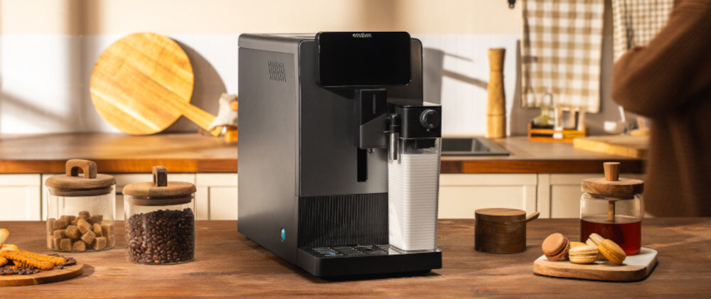 Na drewnianym blacie kuchennym stoi czarny ekspres do kawy z logo \'CECOTEC\' na górze. Wokół znajdują się słoiki z kawą i cukrem oraz talerz z ciastkami.