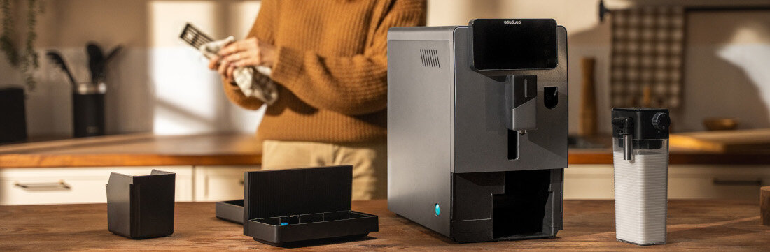 Na drewnianym blacie kuchennym stoi ekspres do kawy z widocznym logo \'CECOTEC\', obok którego znajdują się akcesoria. W tle osoba trzymająca ściereczkę.