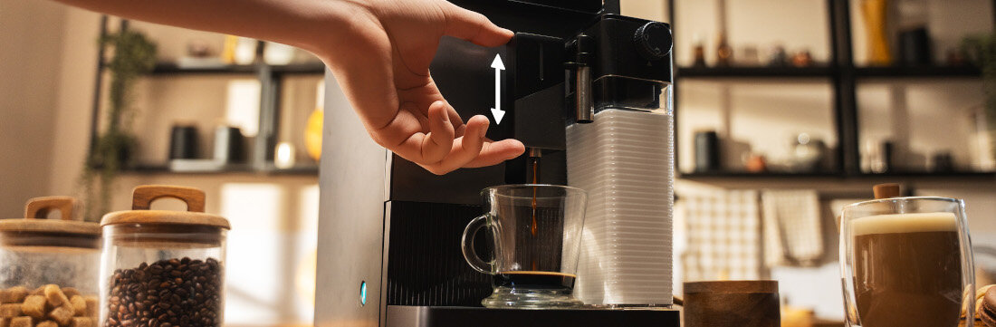 Ekspres do kawy z filiżanką pod dyszą, obok znajdują się słoiki z kawą i cukrem. W tle widać rozmytą kuchnię z półkami i dekoracjami.