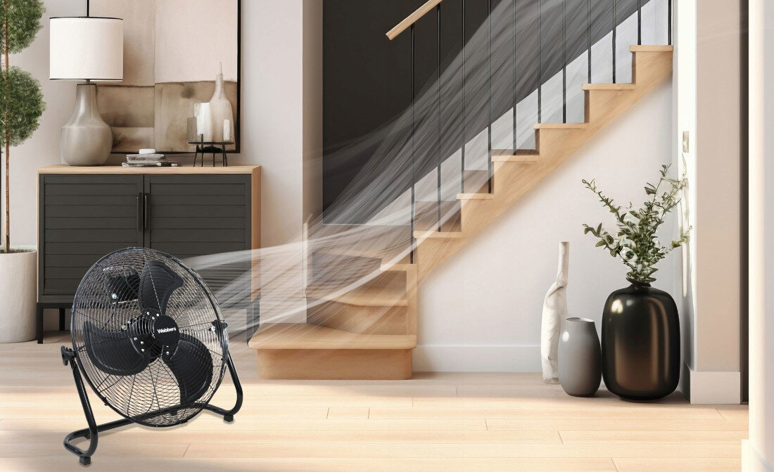 Wentylator stoi na podłodze w nowoczesnym wnętrzu, obok schodów i roślin w wazonach. Na wentylatorze widoczne jest logo \'webber\'.