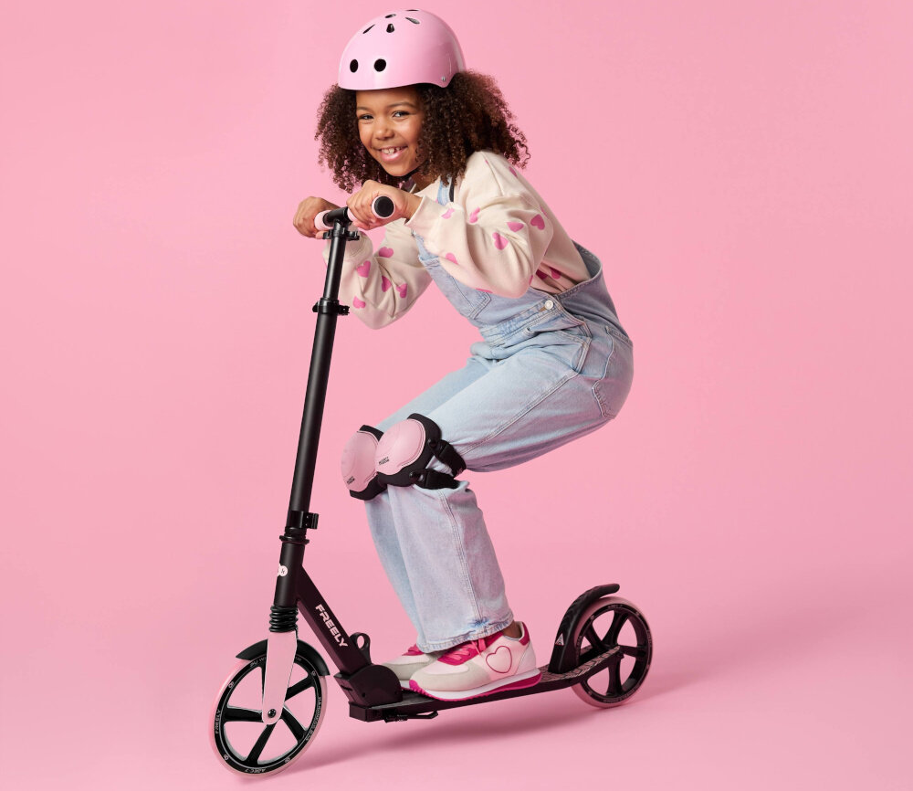 Hulajnoga KINDERKRAFT Freely Różowy różowe tło dziecko na hulanodze w kasku swobodna jazda radność hobby aktywność