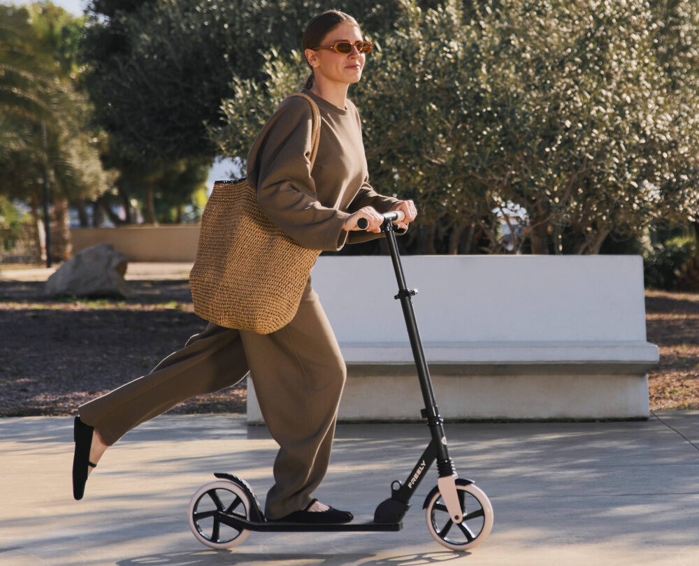 Hulajnoga KINDERKRAFT Freely Beżowy park chodnik osoba jedzie na hulajnodze po drodze w tle drzewa krzewy swobodna jazda radność hobby aktywność