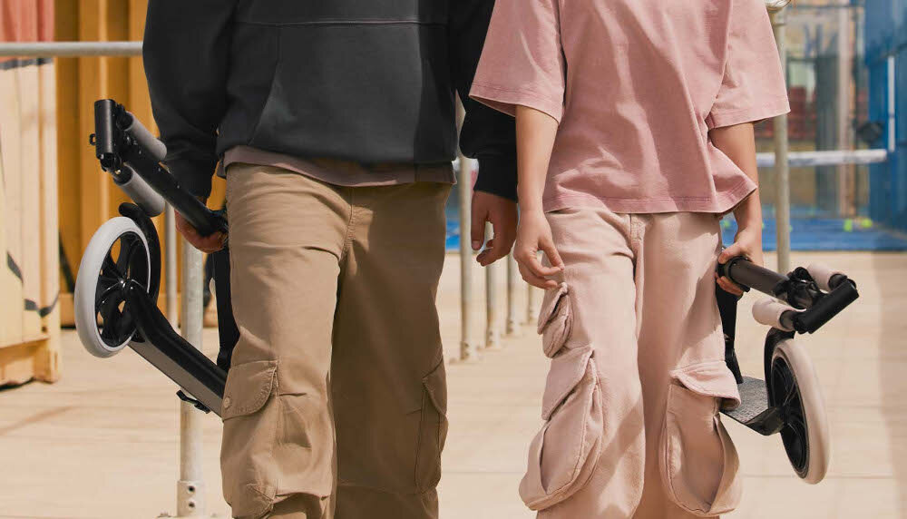 Hulajnoga KINDERKRAFT Freely Beżowy park droga dwie osoby idą po drodze w dłoniach trzymają złożone hulajnogi szybki i intuicyjny mechanizm składania wymiary po złożeniu 82 cm długości 11 cm szerokości 32,5 cm wysokości oszczędzasz miejsce