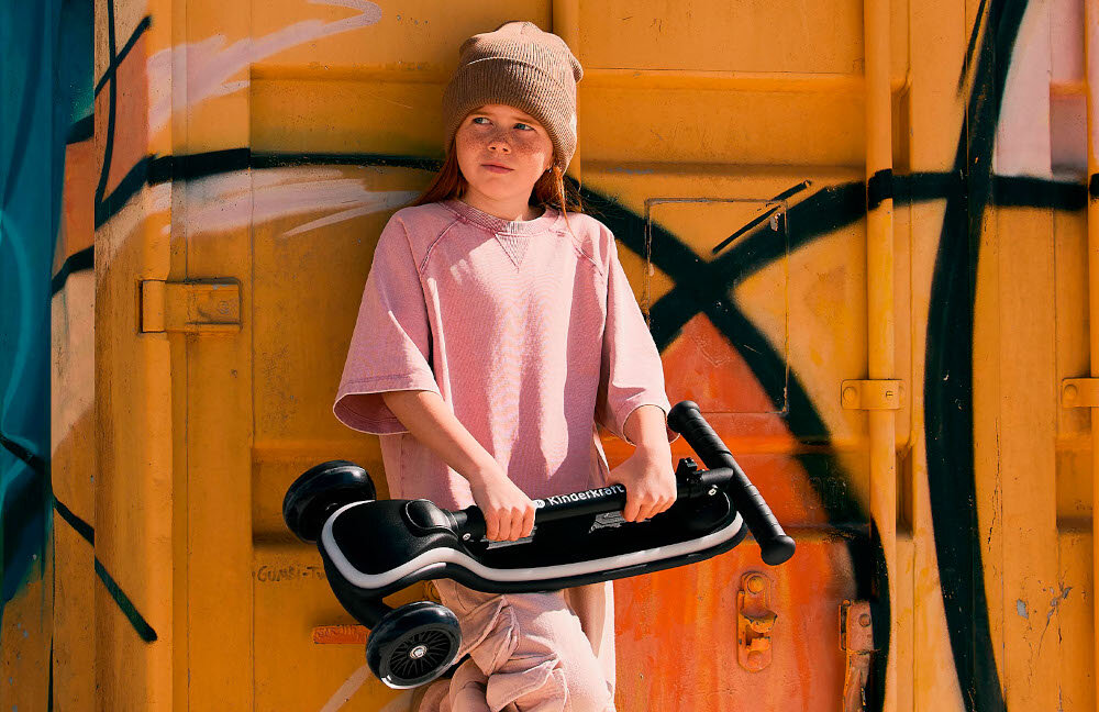 Hulajnoga dla dzieci KINDERKRAFT Raket Beżowy dziecko w czapce opiera się o kolorową ścianę w dłoniach trzyma złożoną hulajnogę waga 3,2 kg łatwa do przenoszenia i składania niewielkie wymiary po złożeniu (15 x 28 x 60 cm) towarzysz codziennych aktywności rodzinnych