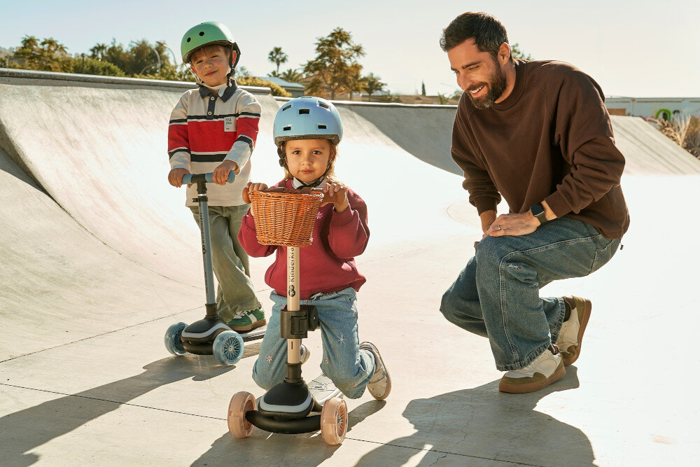 Hulajnoga dla dzieci KINDERKRAFT Raket Up Niebieski skatepark dwoje dzieci jeździ na hulajnogach obok klęczy dorosła osoba w tle drzewa niebo aktywność zabawa hulajnoga 2 w 1