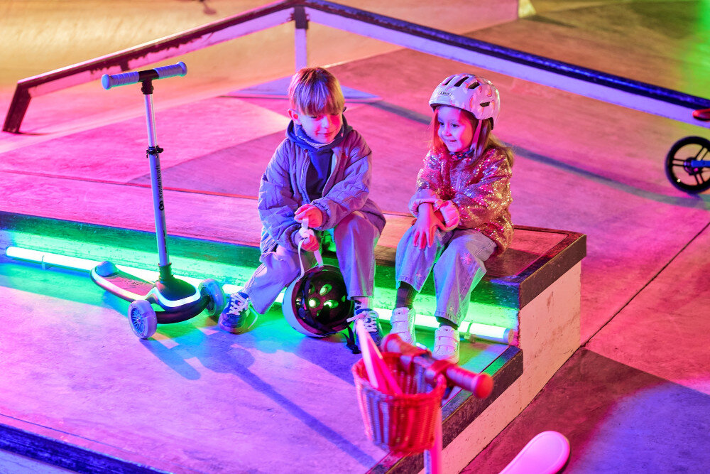 Hulajnoga dla dzieci KINDERKRAFT Raket Up Niebieski skatepark dwoje dzieci siedzi na rampie obok stoją hulajnogi kolorowe światła spełnia europejskie normy bezpieczeństwa EN wysoka jakość wykonania konstrukcja została przetestowana pod kątem trwałości, stabilności oraz bezpieczeństwa użytkowania sprzęt dopasowany do obowiązujących standardów