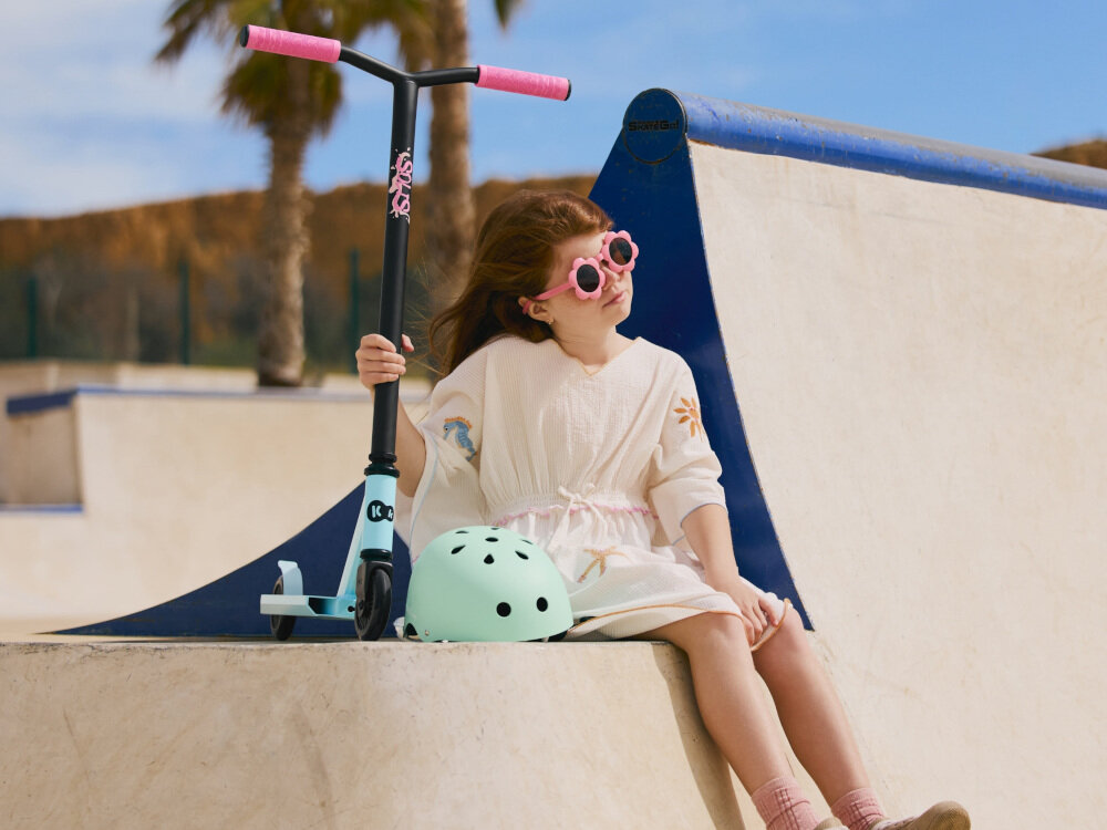 Hulajnoga wyczynowa KINDERKRAFT Solis Różowy skatepark osoba rampie skatepark opiera dłoń na hulajnodze w tle błękitne niebo palma dla młodszych i starszych aktywność hobby rozwój zabawa