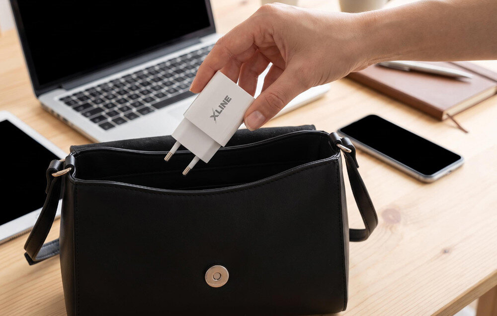 Osoba wkłada białą ładowarkę z logo \'XLINE\' do czarnej torebki. W tle znajduje się otwarty laptop, smartfon i notatnik na drewnianym stole.