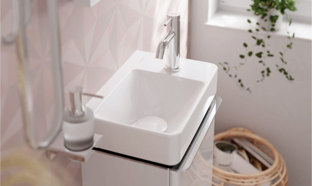 BATERIA UMYWALKOWA HANSGROHE TECTURIS Zbliżenie na nablatową umywalkę w kolorze białym z chromowaną baterią. Obok znajduje się dozownik mydła, a w tle widoczna jest jasna łazienka z rośliną i koszem na ręczniki, co podkreśla minimalistyczny i funkcjonalny styl wnętrza