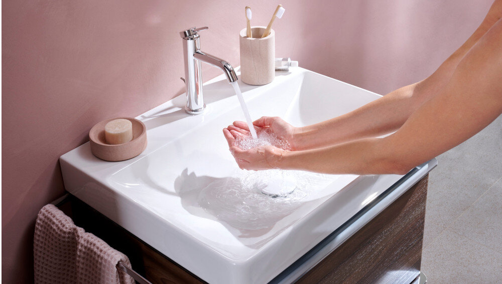BATERIA UMYWALKOWA HANSGROHE TECTURIS Bateria umywalkowa zamontowana przy ceramicznej umywalce z płynącym strumieniem wody. Obok znajdują się mydelniczka, kubek na szczoteczki i ręcznik w dopasowanej kolorystyce