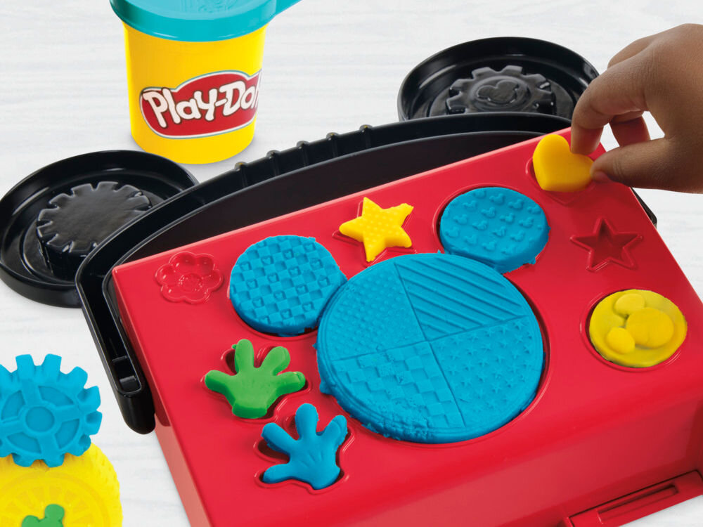 Ciastolina PLAY-DOH Disney Junior Mickey Mouse Przenośny garaż Dziecko wyciska kształty w czerwonej walizeczce Play-Doh, która służy jako matryca. Na powierzchni widoczne odciśnięte wzory w niebieskiej i zielonej ciastolinie, w tym charakterystyczna głowa Myszki Miki z teksturami, gwiazdka oraz rączka.