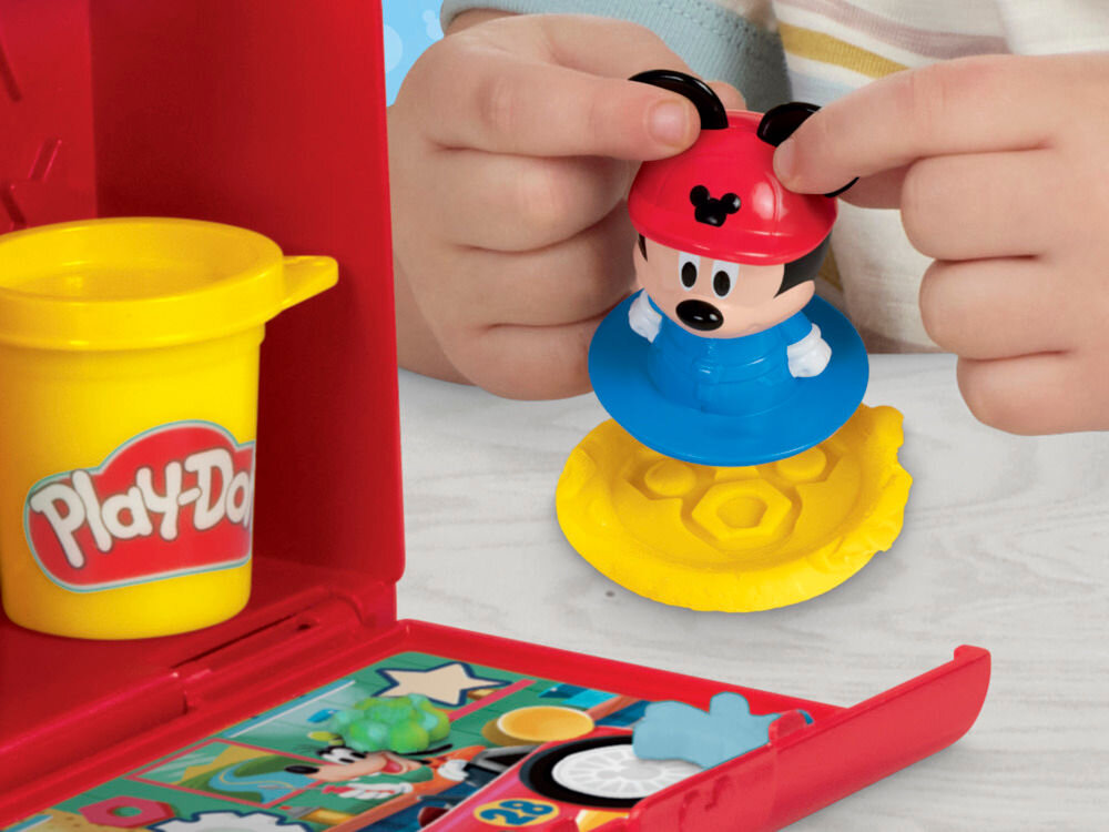 Ciastolina PLAY-DOH Disney Junior Mickey Mouse Przenośny garaż Zbliżenie na figurkę Myszki Miki w stroju mechanika, którą dziecko odciska wzór w żółtej masie plastycznej Play-Doh. W tle widoczna otwarta czerwona walizeczka z kolorowymi ilustracjami Disneya.