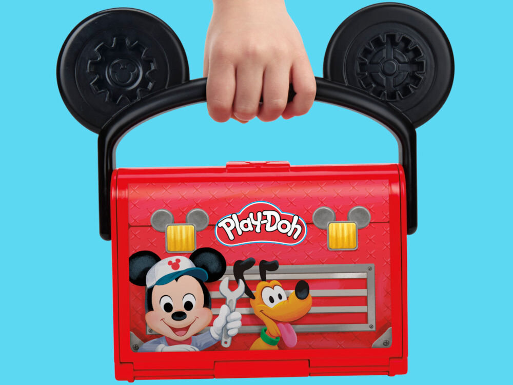 Ciastolina PLAY-DOH Disney Junior Mickey Mouse Przenośny garaż Dziecko trzyma za czarną rączkę w kształcie uszu Myszki Miki zamkniętą czerwoną walizeczkę Play-Doh. Na froncie walizki znajduje się kolorowa grafika z Myszką Miki i psem Pluto w warsztacie.